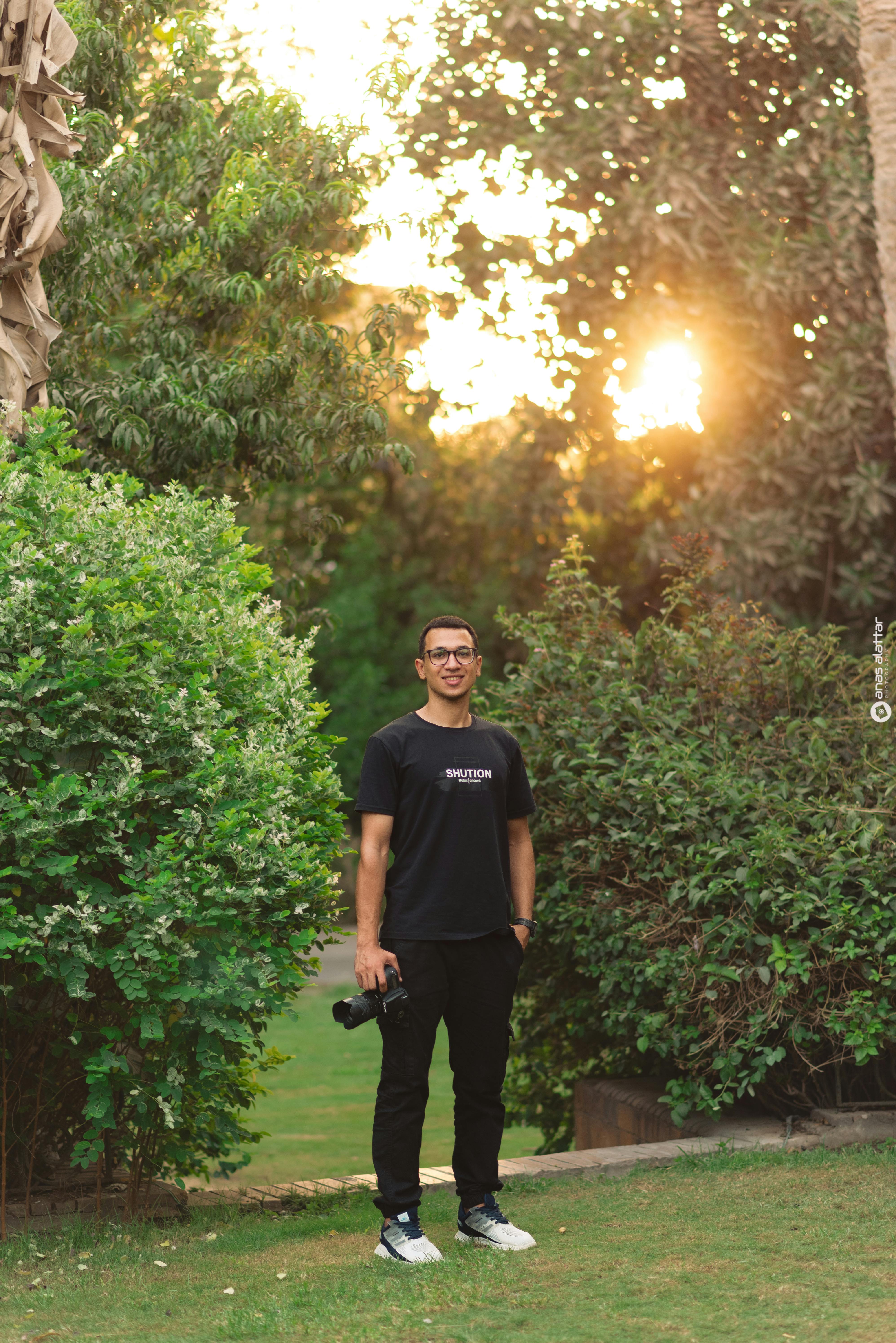 De franc Un fotògraf es troba amb confiança en un jardí verd durant la posta de sol, sostenint una càmera. Foto d'estoc