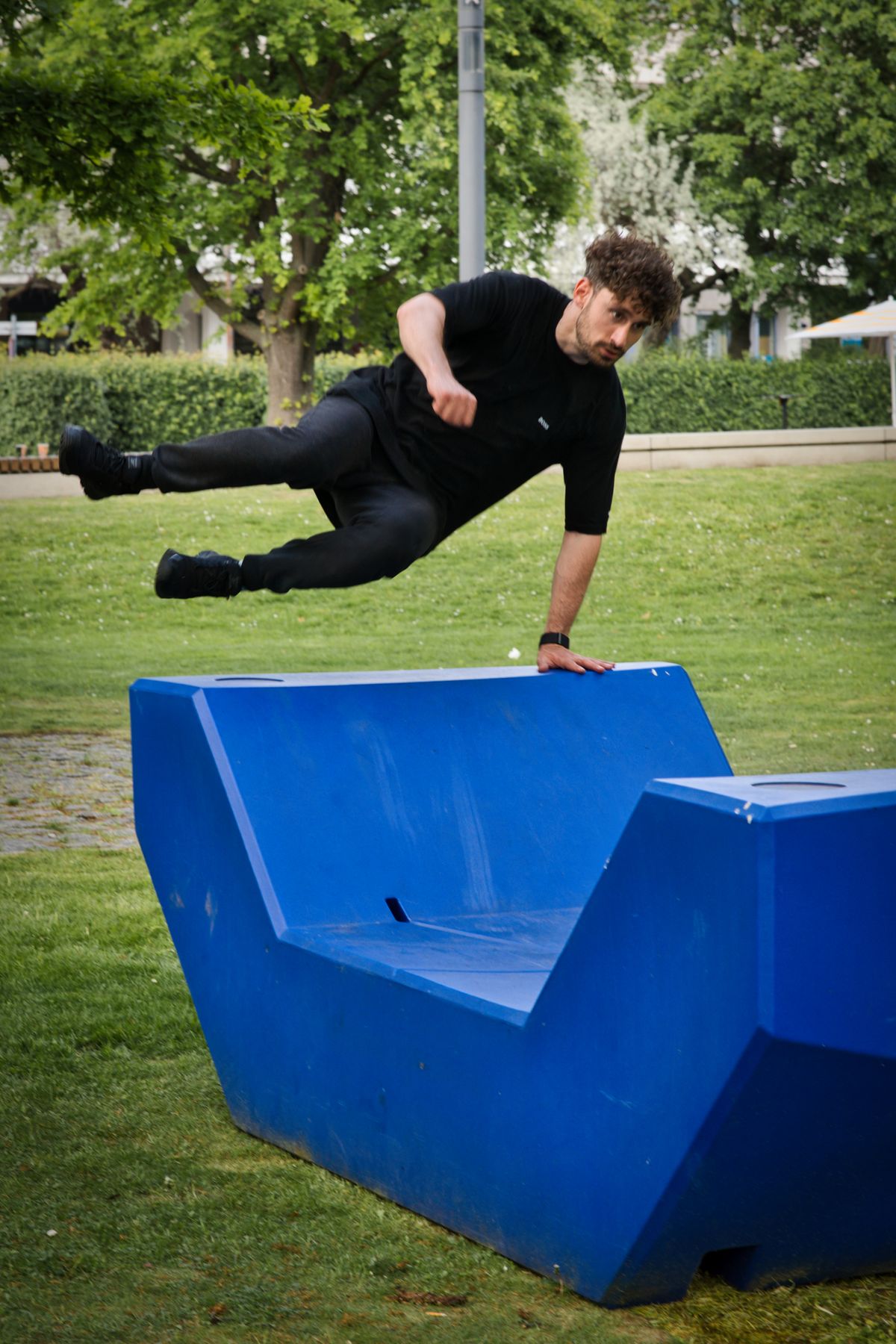 A traceur vaulting a purpose-built blue parkour obstacle set on grass in a public park.