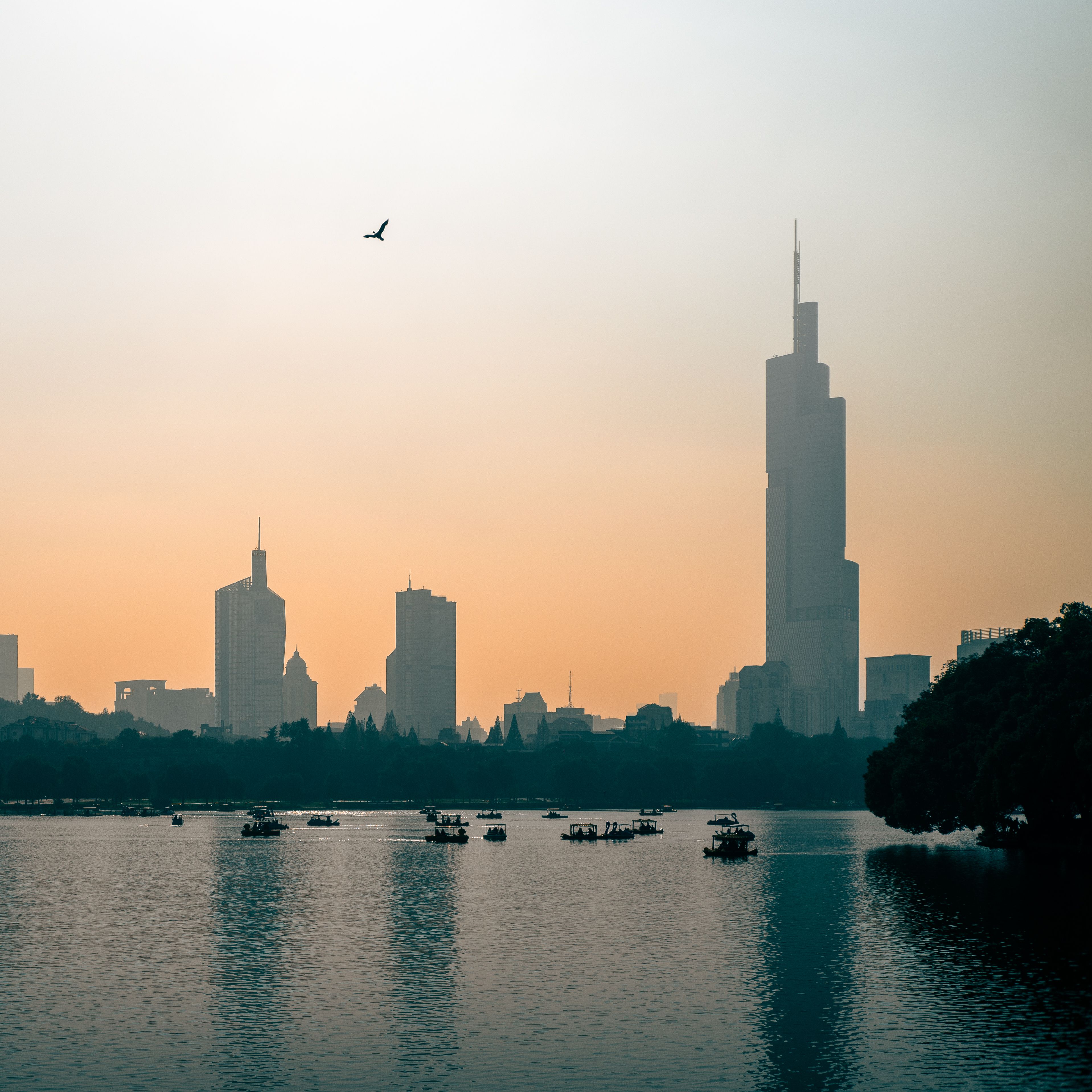 Siluet cakrawala kota Nanjing berlatar belakang matahari terbenam yang hangat, dengan Danau Xuanwu dan perahu di latar depan. Via Pexels (by Cheng Shi Song)