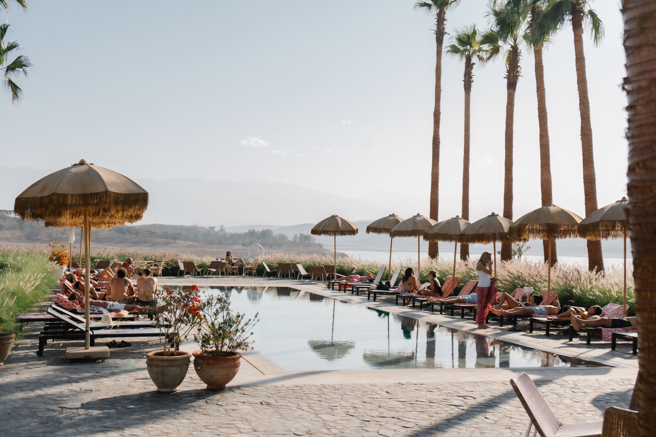 People relaxing by a luxurious outdoor pool with palm trees and parasols under a sunny sky.