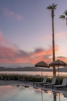 Tranquil poolside view at sunset with palm trees and umbrellas reflecting in the water.