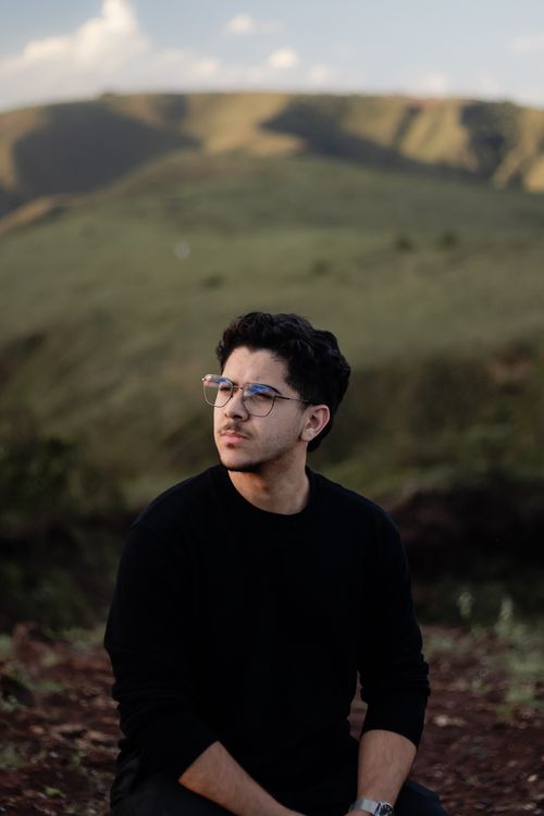 Free Young man with glasses outdoors, relaxed expression against a green hills background. Stock Photo