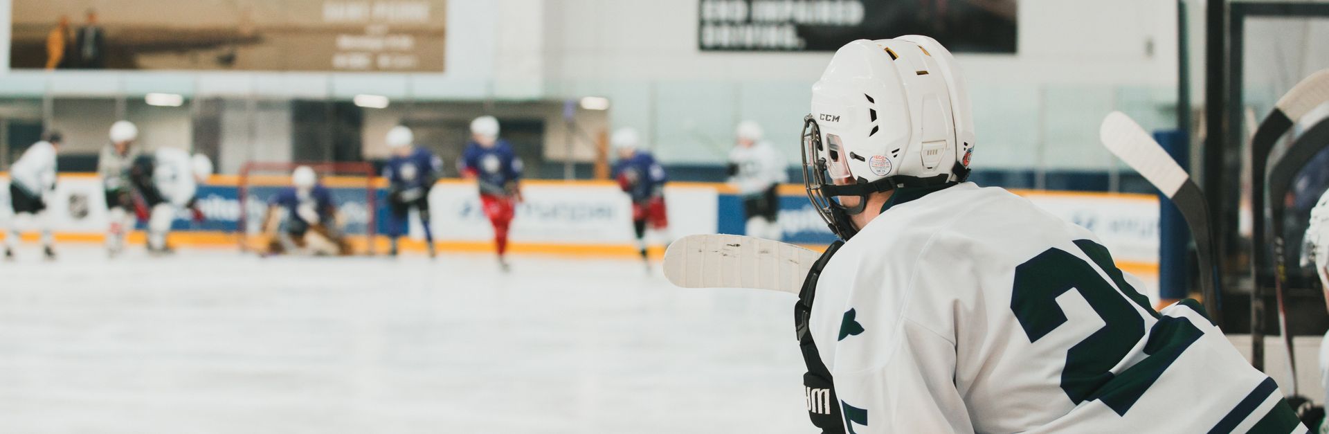 Adult ice hockey player in white jersey observing match from bench indoors.