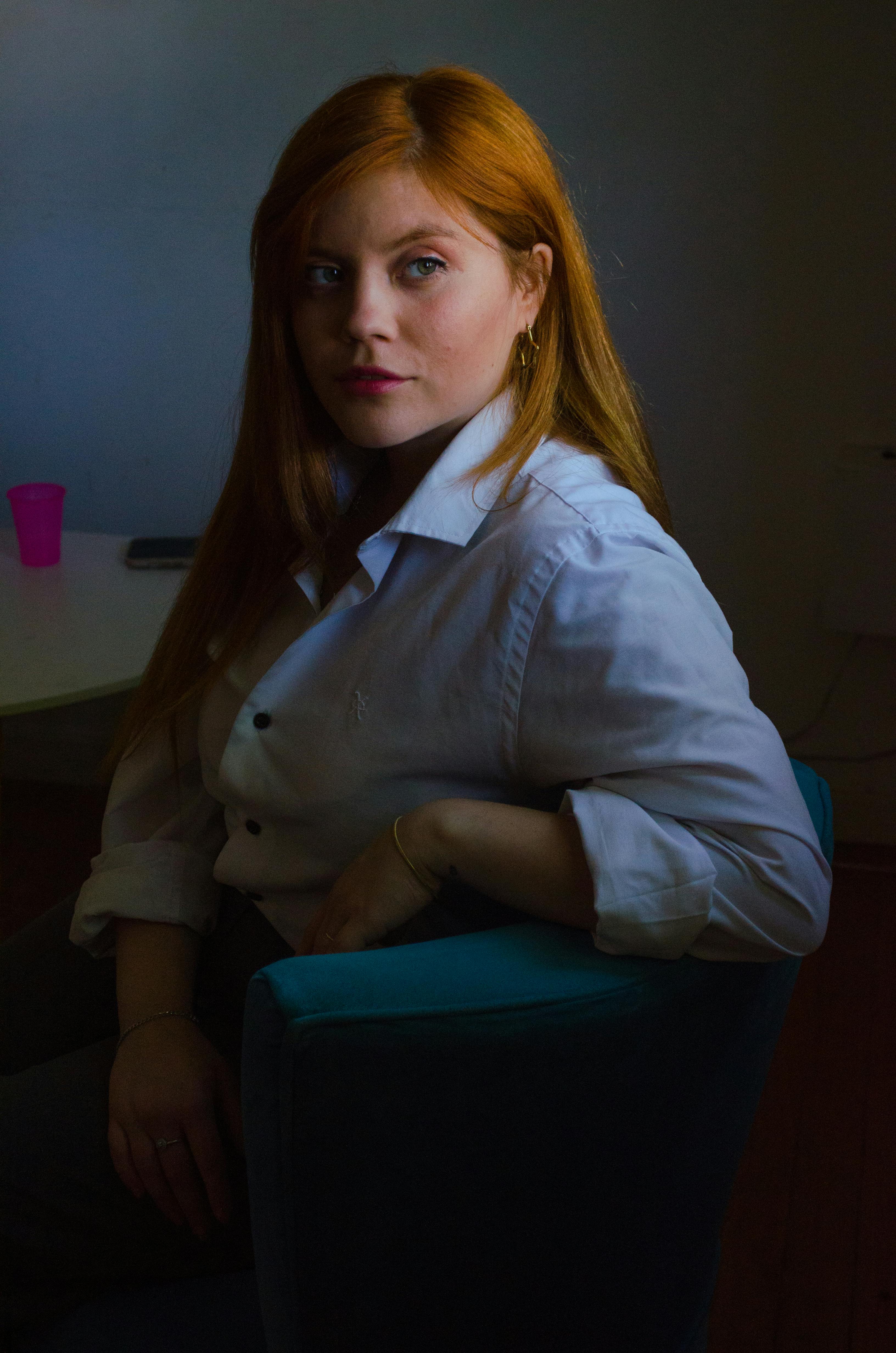 gratis Portret van een roodharige vrouw in een wit shirt, zittend binnen met een peinzende blik. Stockfoto