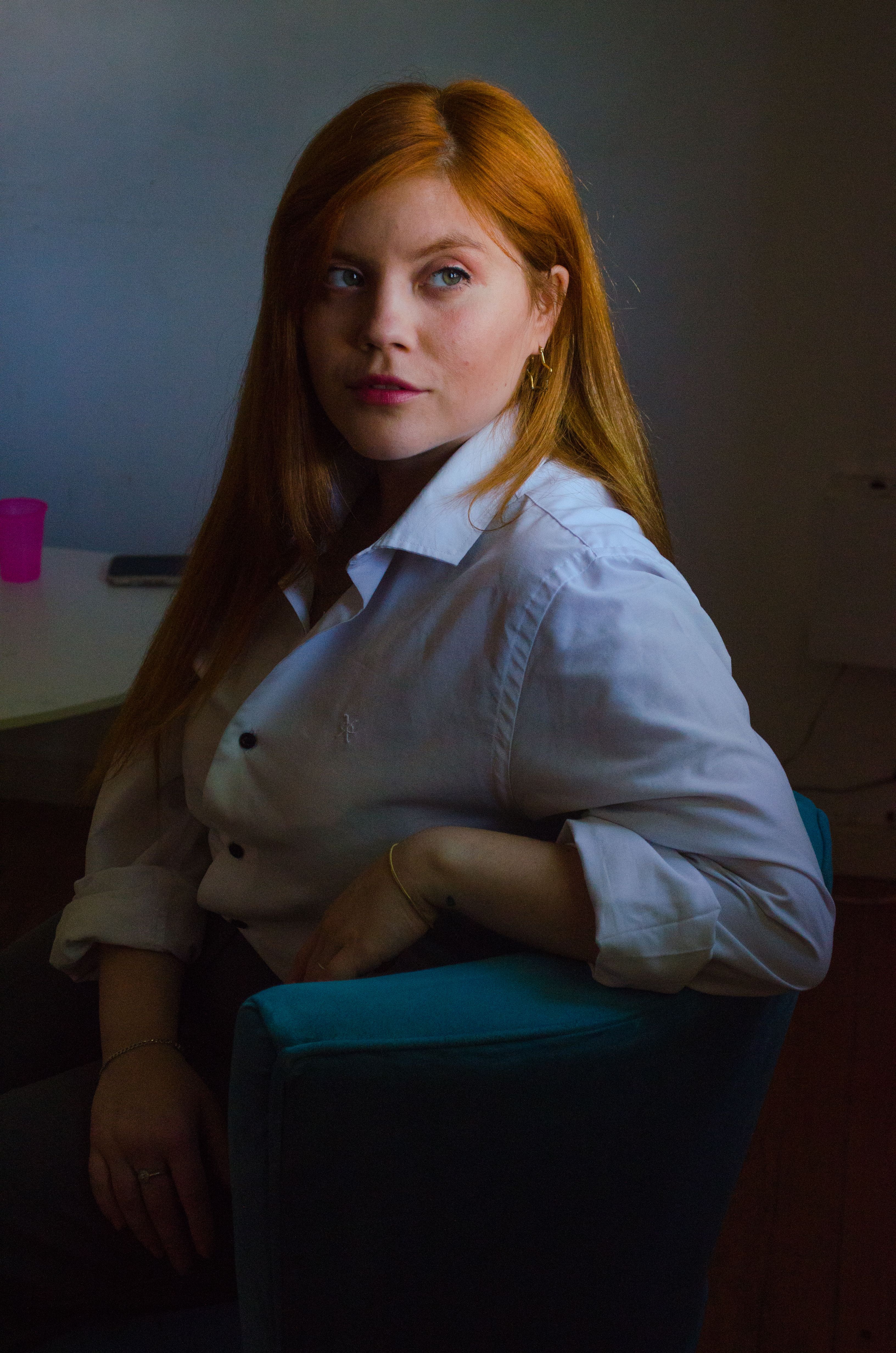 Contemplative woman with red hair in a white shirt sitting indoors with soft natural lighting.