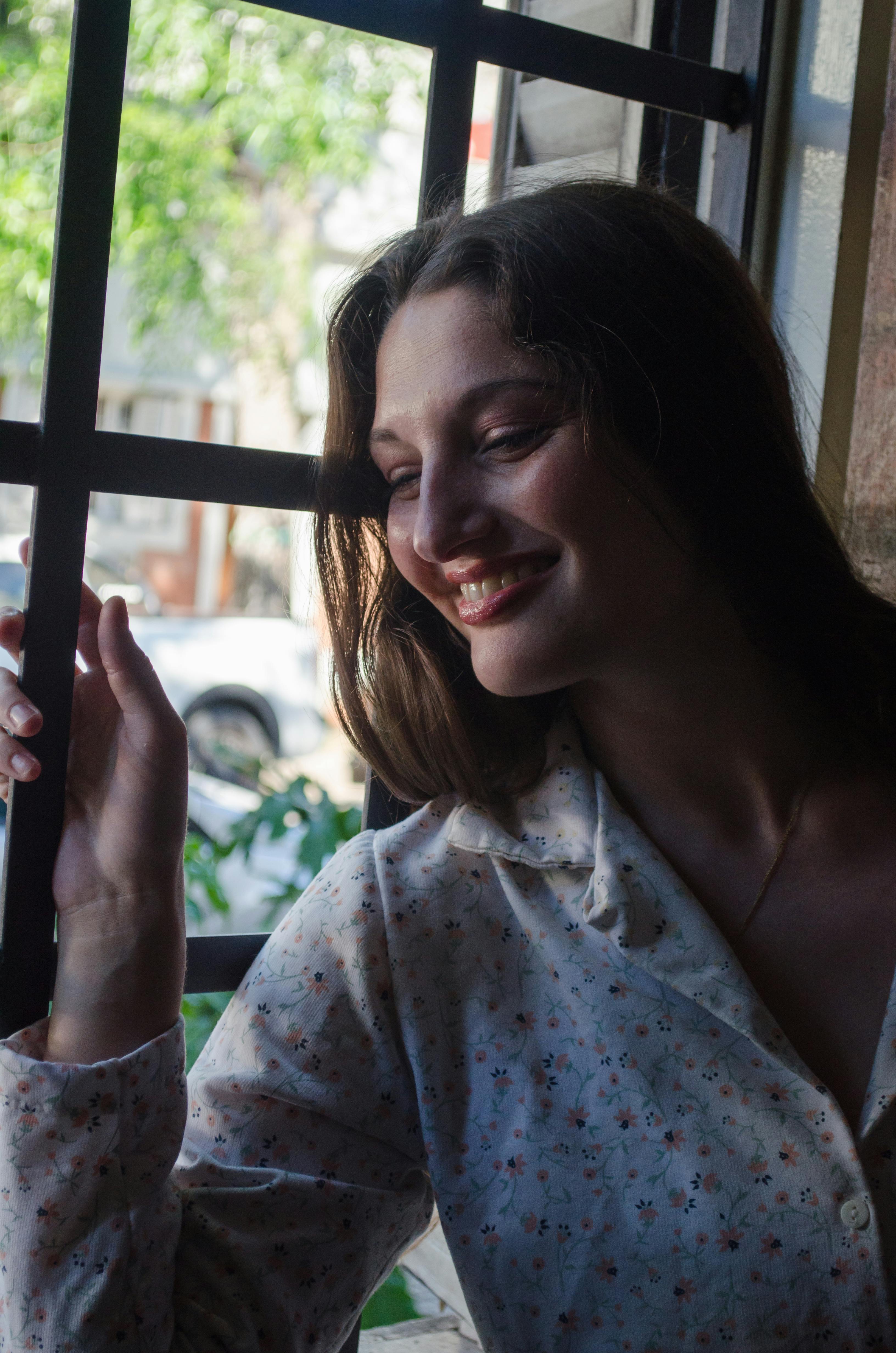 무료 한 여성이 실내에서 부드러운 자연광이 들어오는 햇살이 비치는 창문 옆에 서서 평화롭게 미소 짓고 있습니다. 스톡 사진