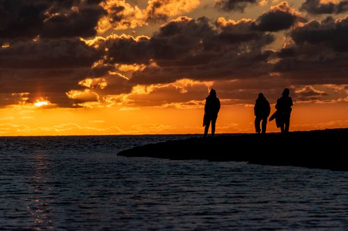 Free Three people walk along the ocean's edge, silhouetted against a vibrant sunset. Stock Photo