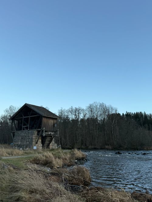 Free A rustic wooden mill stands by a tranquil river, surrounded by bare trees in a serene landscape. Stock Photo