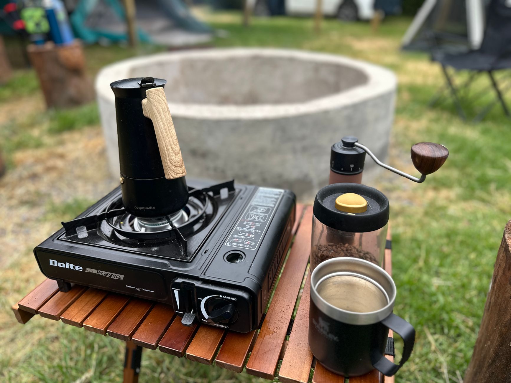 Outdoor coffee setup with a portable stove, kettle, and coffee grinder near a fire pit.