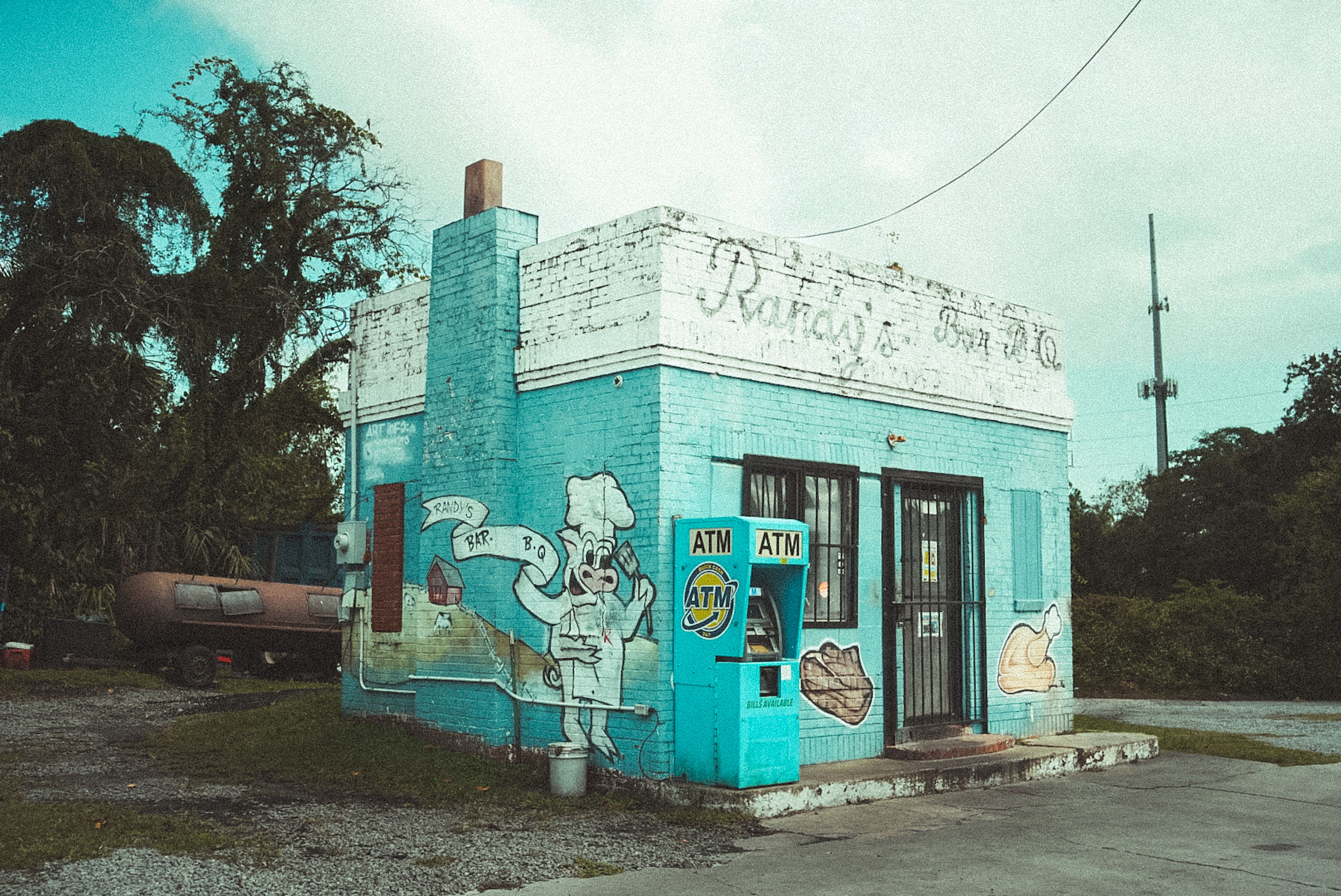 A retro blue BBQ building with vibrant murals and an ATM, set against a lush backdrop.