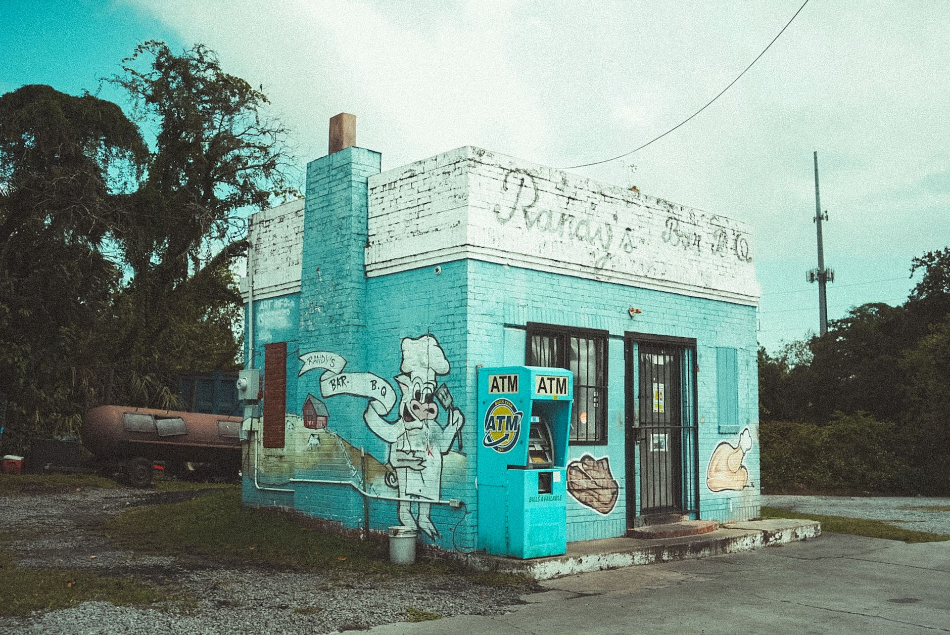 A retro blue BBQ building with vibrant murals and an ATM, set against a lush backdrop.