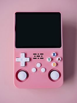 A vintage handheld gaming console in pink with colorful buttons set against a pastel pink background.