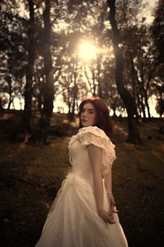 A woman in a vintage white dress stands in a sunlit forest, evoking a mystical bridal scene.