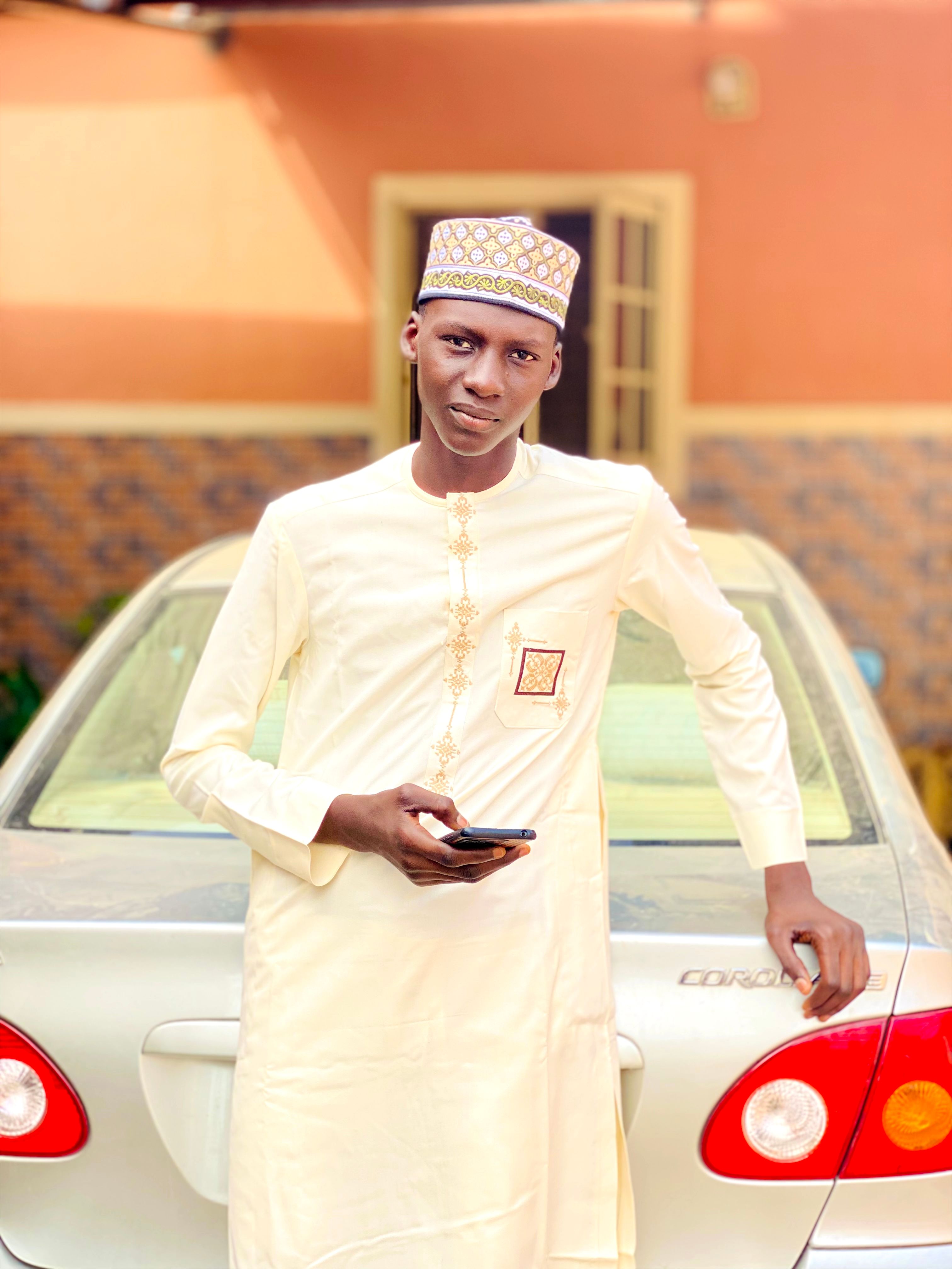 Young man in traditional dress leaning on car holding phone.