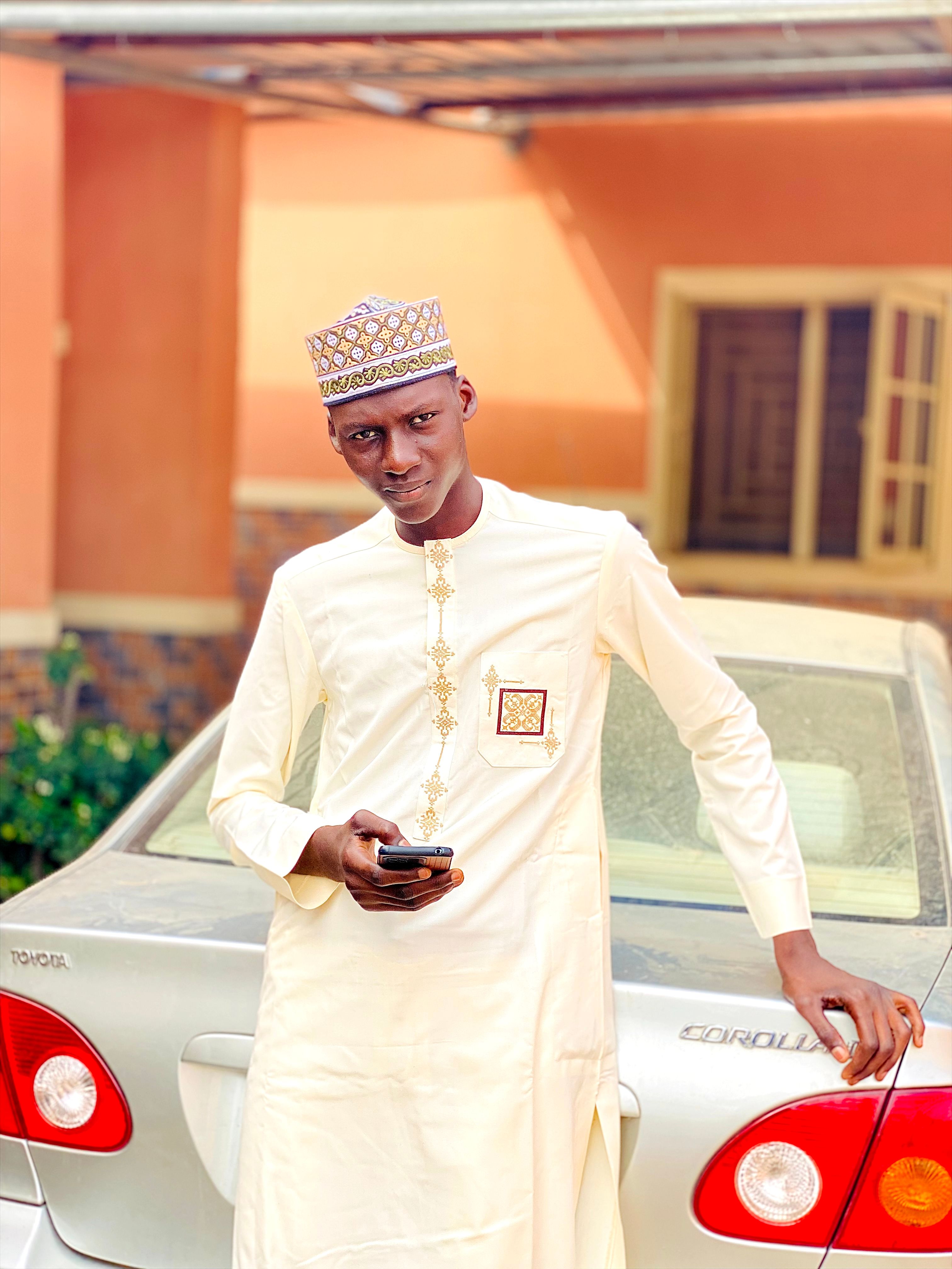A young man in traditional attire poses confidently beside a car outdoors.