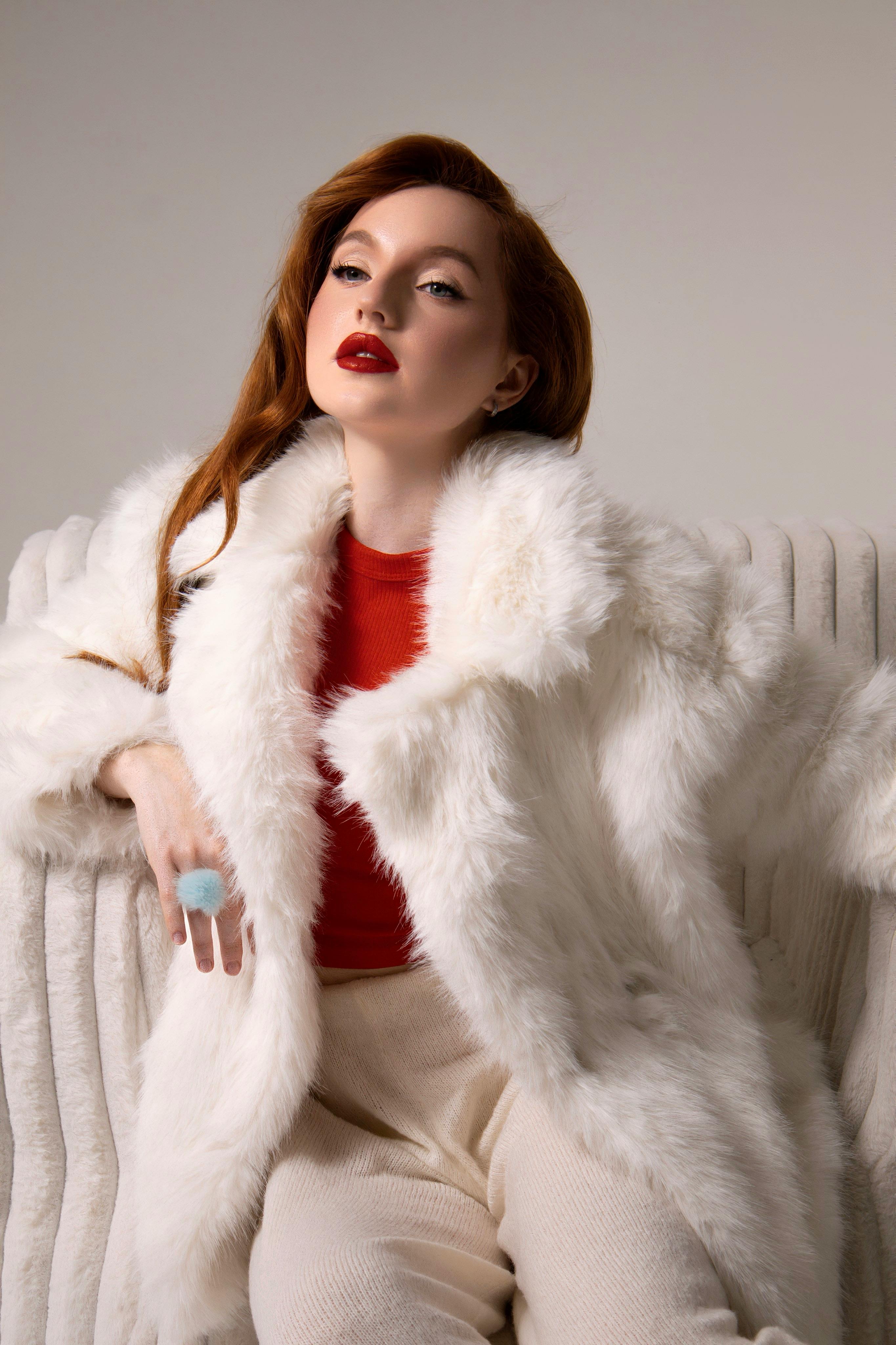 Stylish woman in luxurious white fur coat seated indoors with red lipstick and blouse.