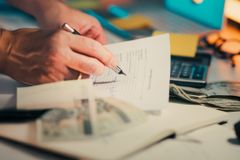 A hand signing financial documents on a desk with cash and a calculator, signifying finance management.