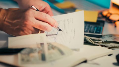 A hand signing financial documents on a desk with cash and a calculator, signifying finance management.