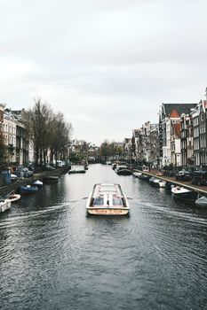 A picturesque view of a boat navigating through Amsterdam's historic canal on a cloudy day.