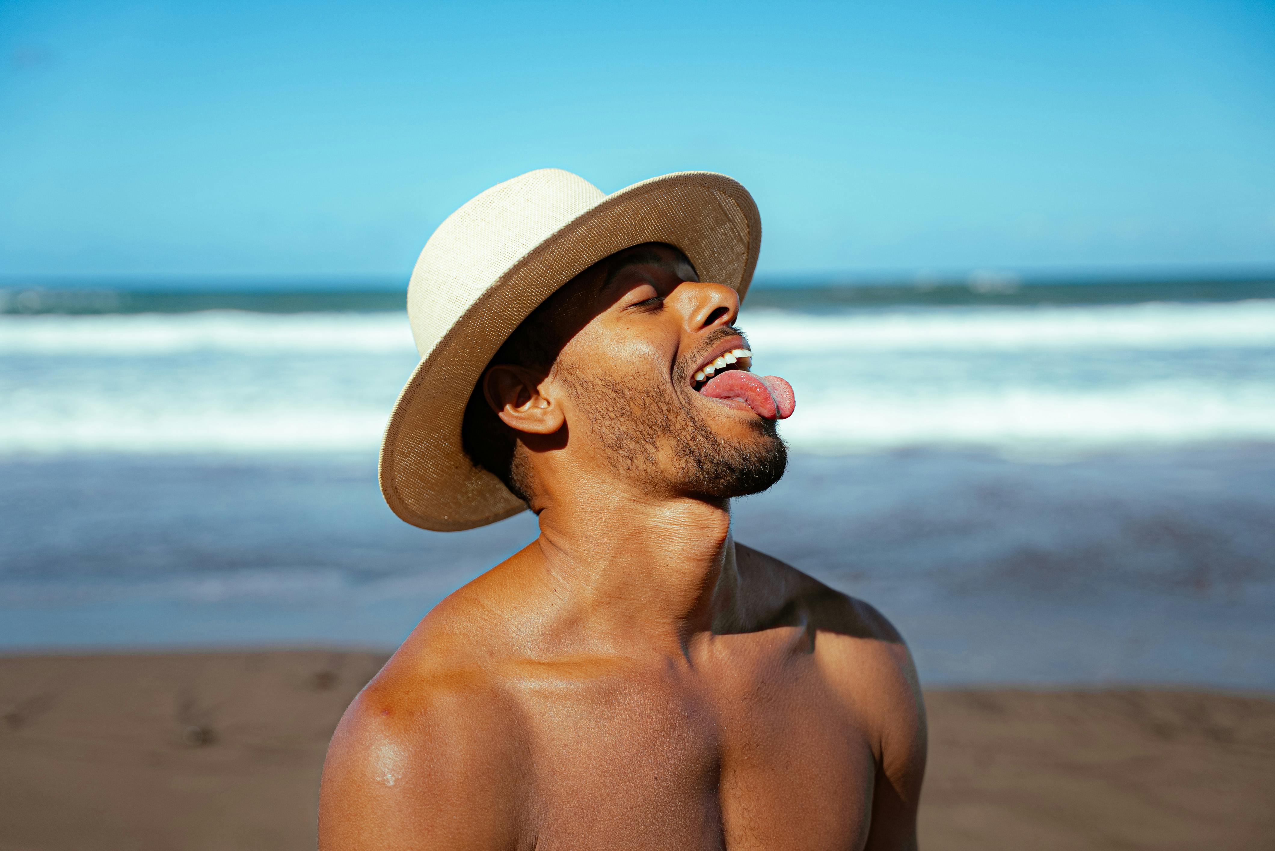 Man in a straw hat enjoying a sunny day at the beach, tongue out, against a blue sky backdrop on Canary Islands.