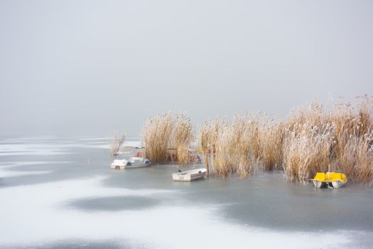 A serene winter landscape featuring frozen lake waters, moored boats, and snow-covered reeds.