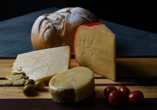 A selection of cheeses and rustic bread on a wooden board with olives and tomatoes.
