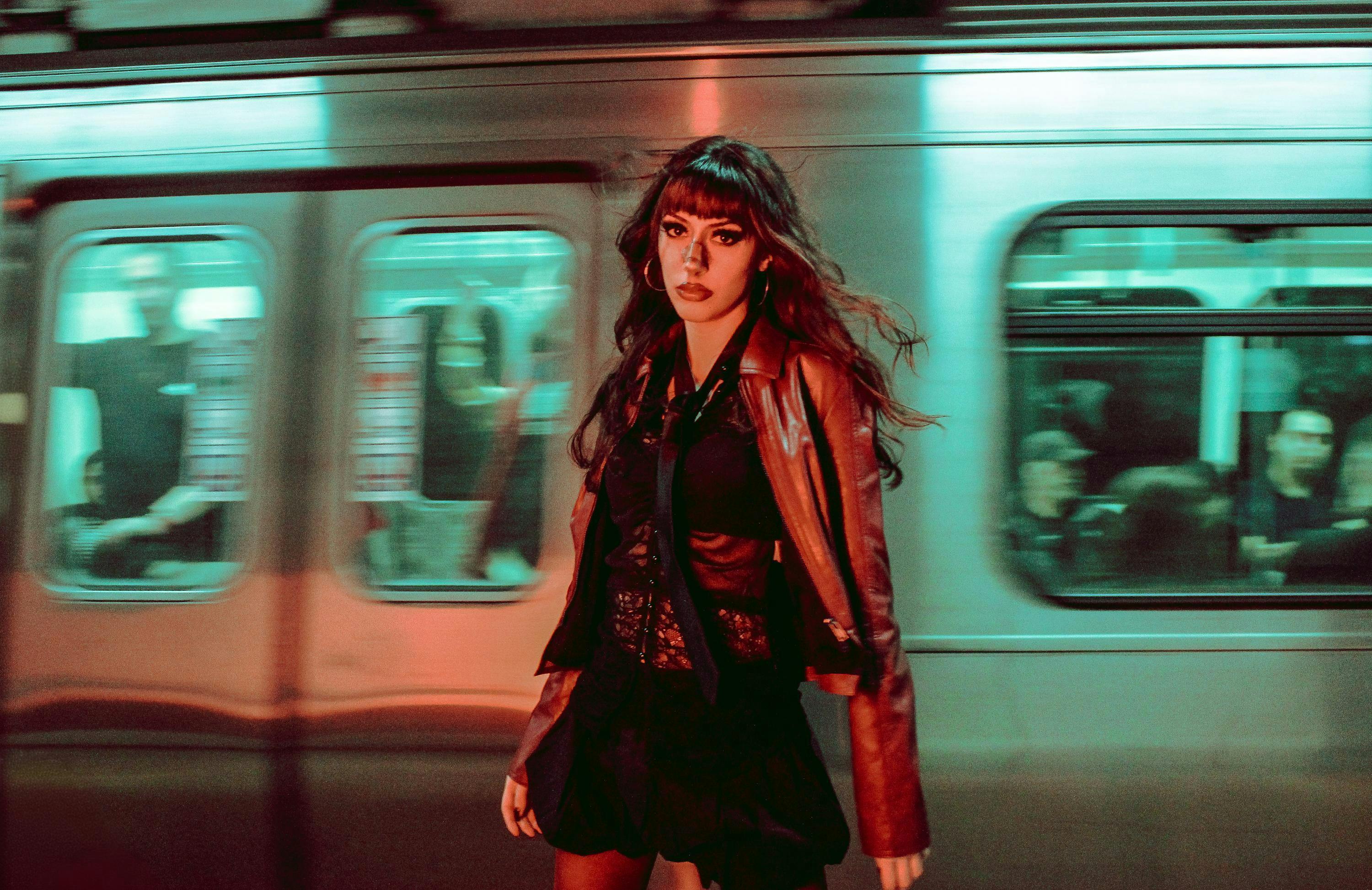 Stylish woman in a leather jacket in front of a moving subway train at night.