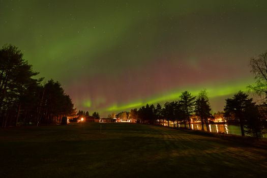 The Northern Lights illuminate the night sky above a tranquil lakeside landscape with a forest backdrop.