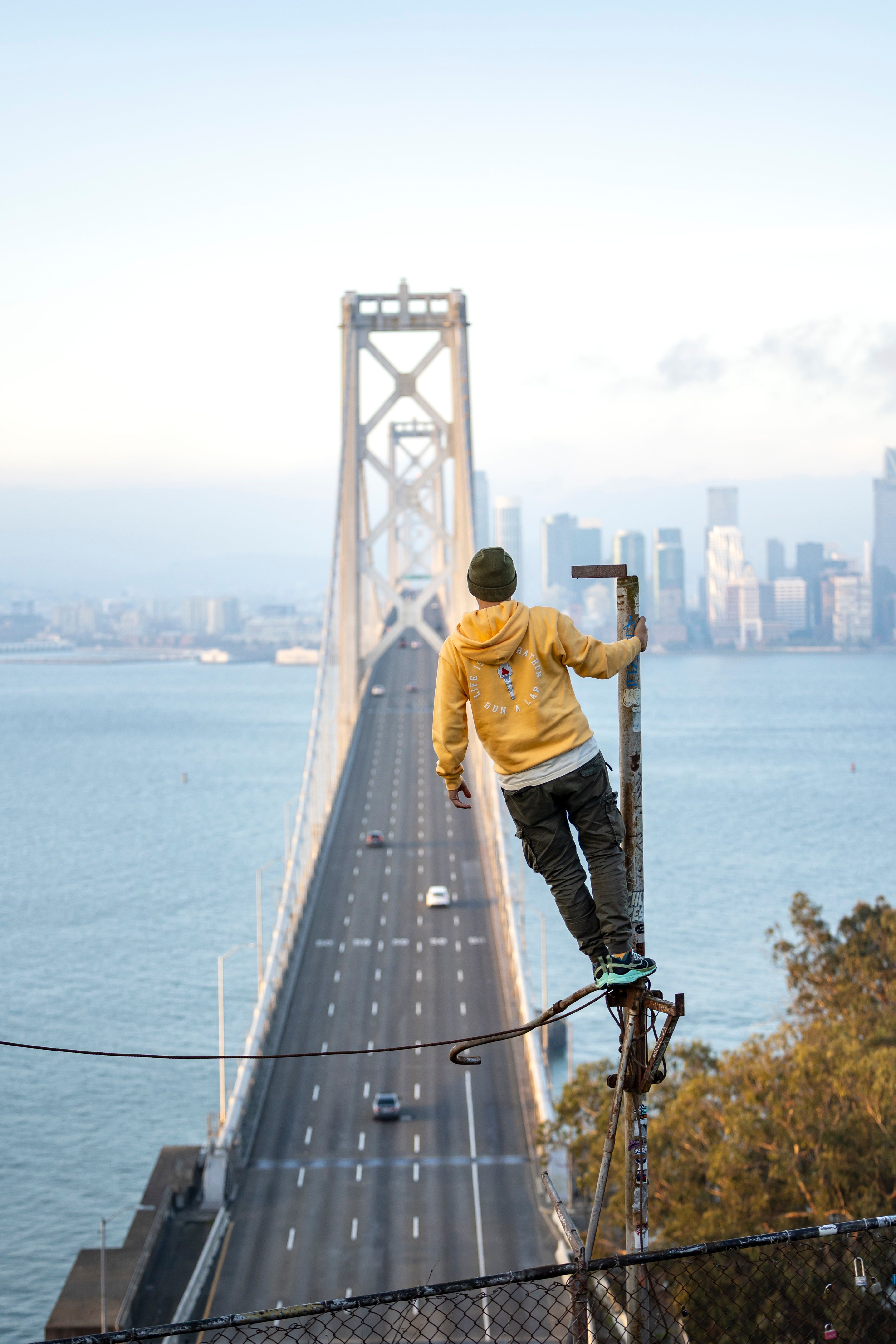 Adventurer standing atop pole with San Francisco's Bay Bridge and skyline backdrop.