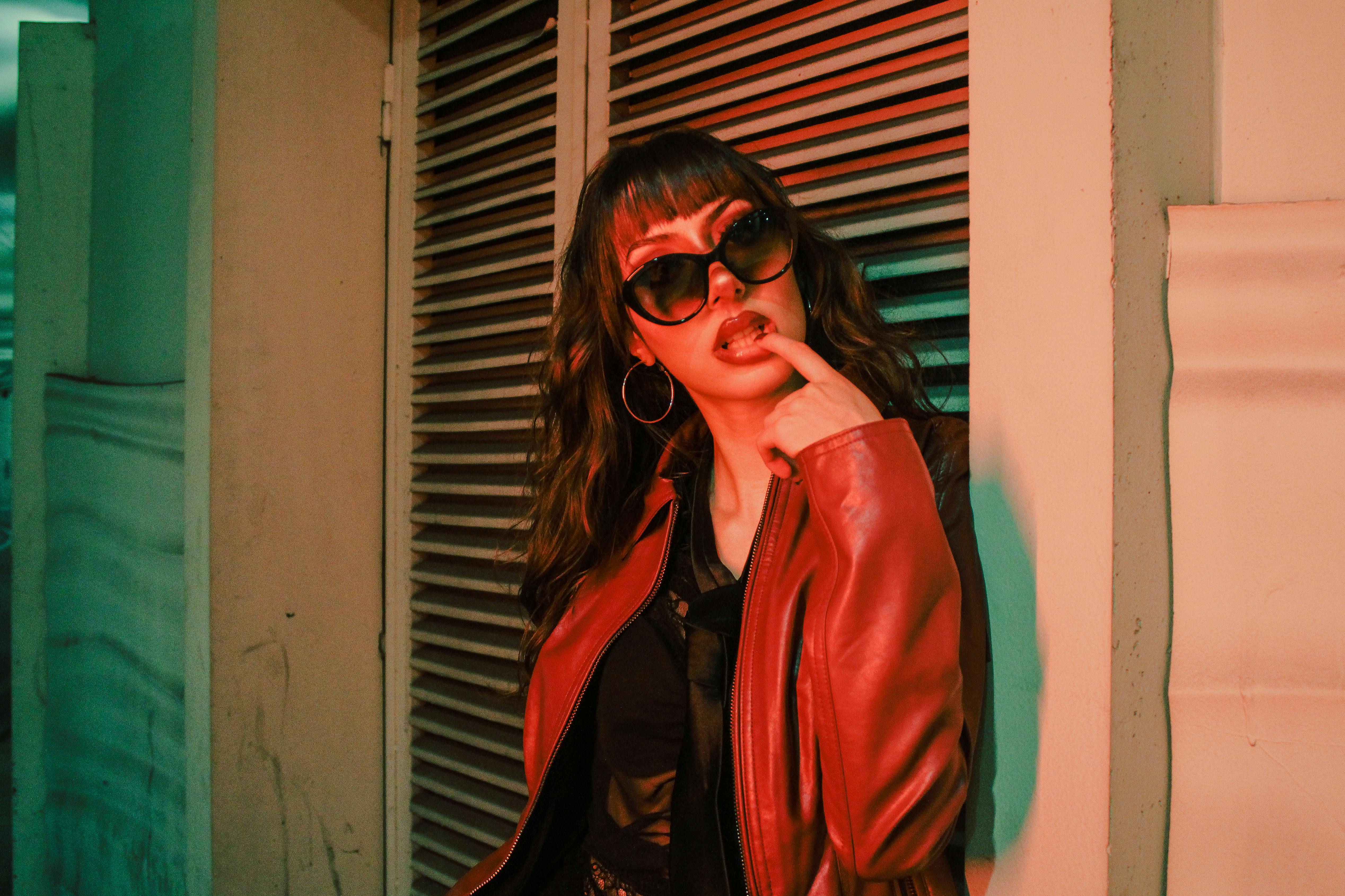 Trendy young woman in a red jacket and sunglasses poses confidently against an urban backdrop.