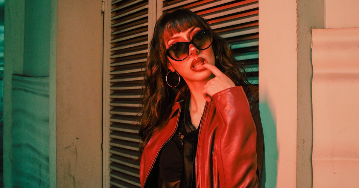 Trendy young woman in a red jacket and sunglasses poses confidently against an urban backdrop.