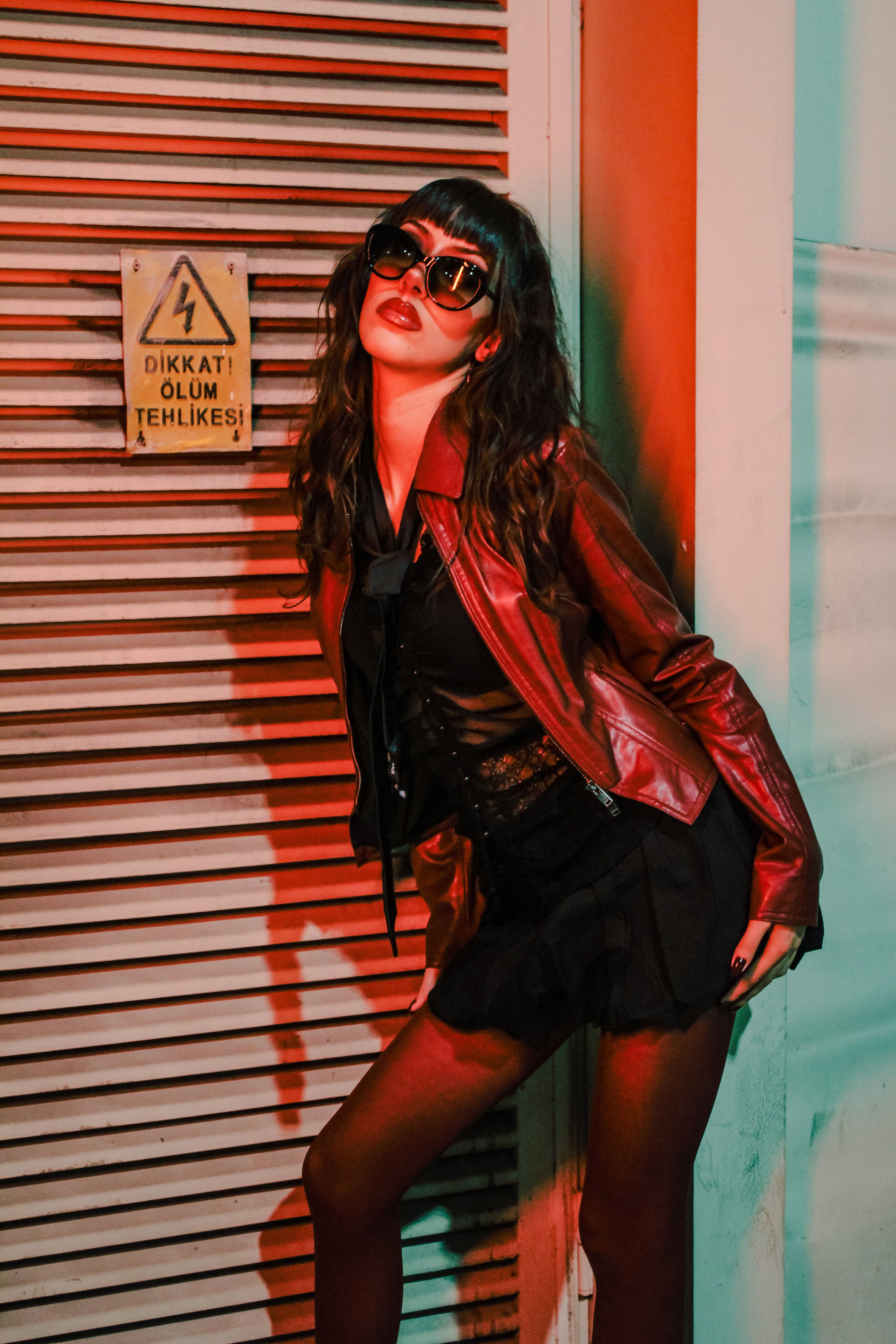 Stylish woman in sunglasses and leather jacket posing against urban backdrop with vibrant neon lighting.