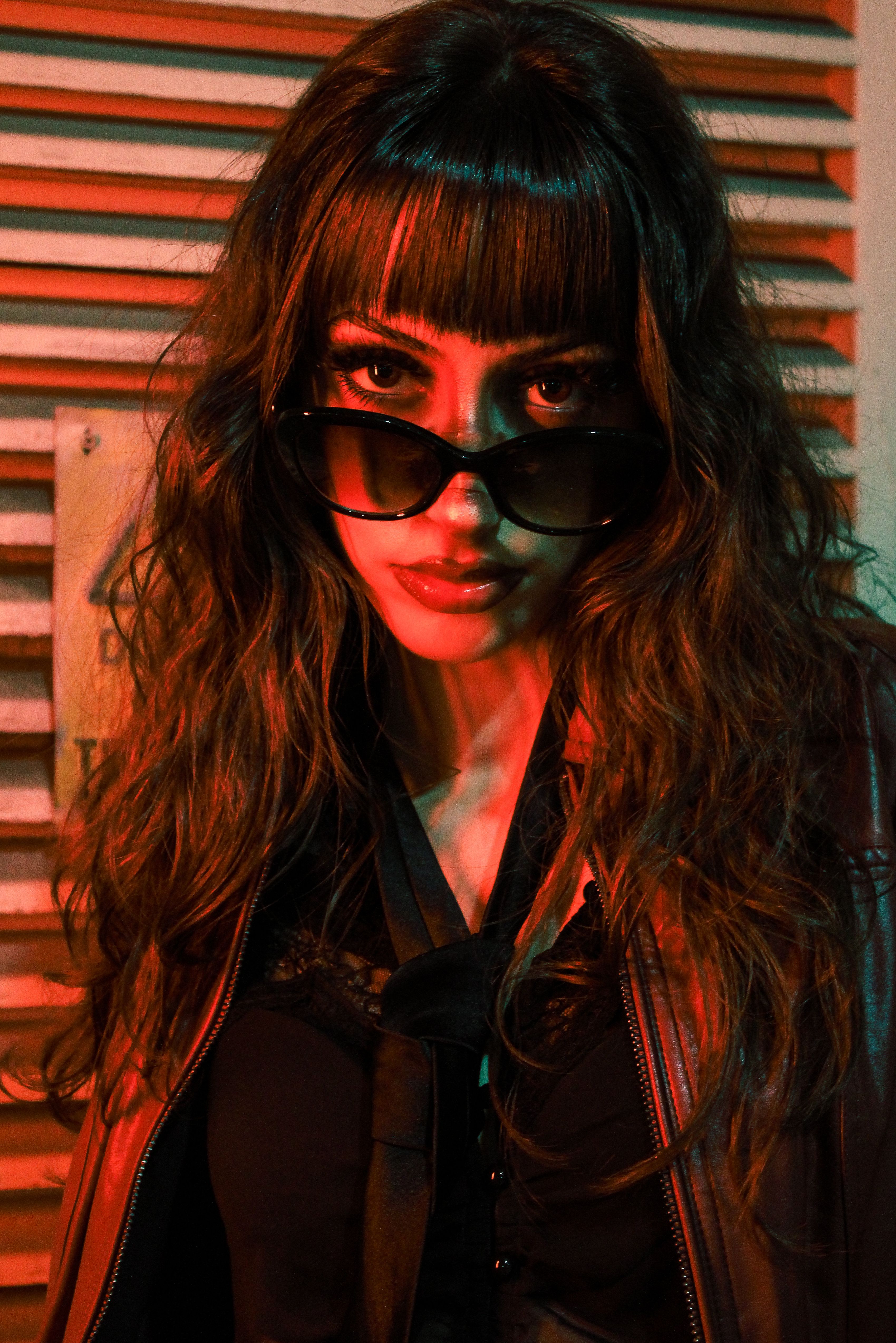 Stylish woman in sunglasses and leather jacket in dramatic red lighting.