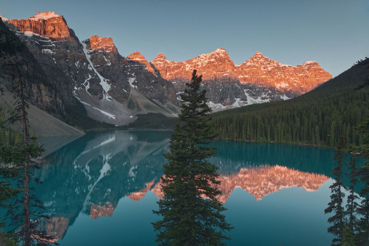 Moraine Lake at sunrise, the Valley of the Ten Peaks glowing red-orange above turquoise water with a near-perfect mirror reflection, pine trees framing the shore.