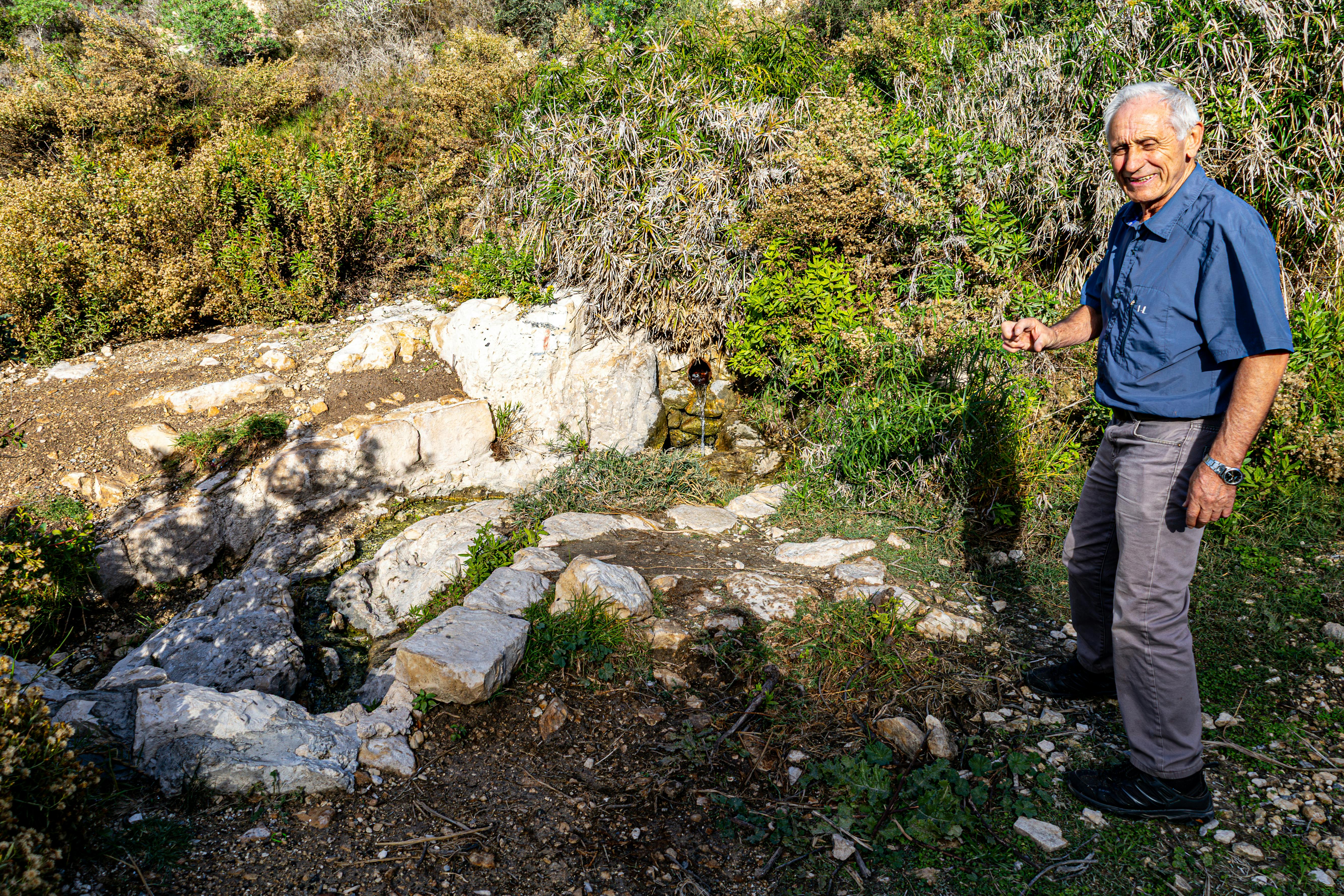 A joyful senior man poses by a natural spring in a rocky outdoor setting under bright sunlight.