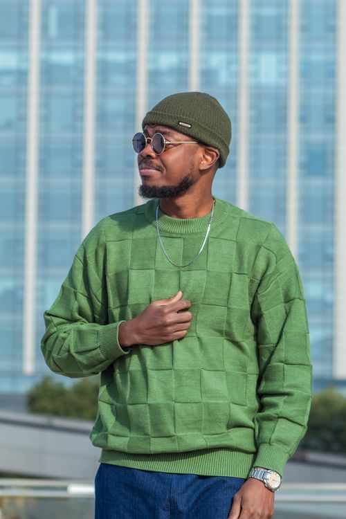 Free Stylish man in green sweater and beanie standing outdoors with a city building backdrop. Stock Photo
