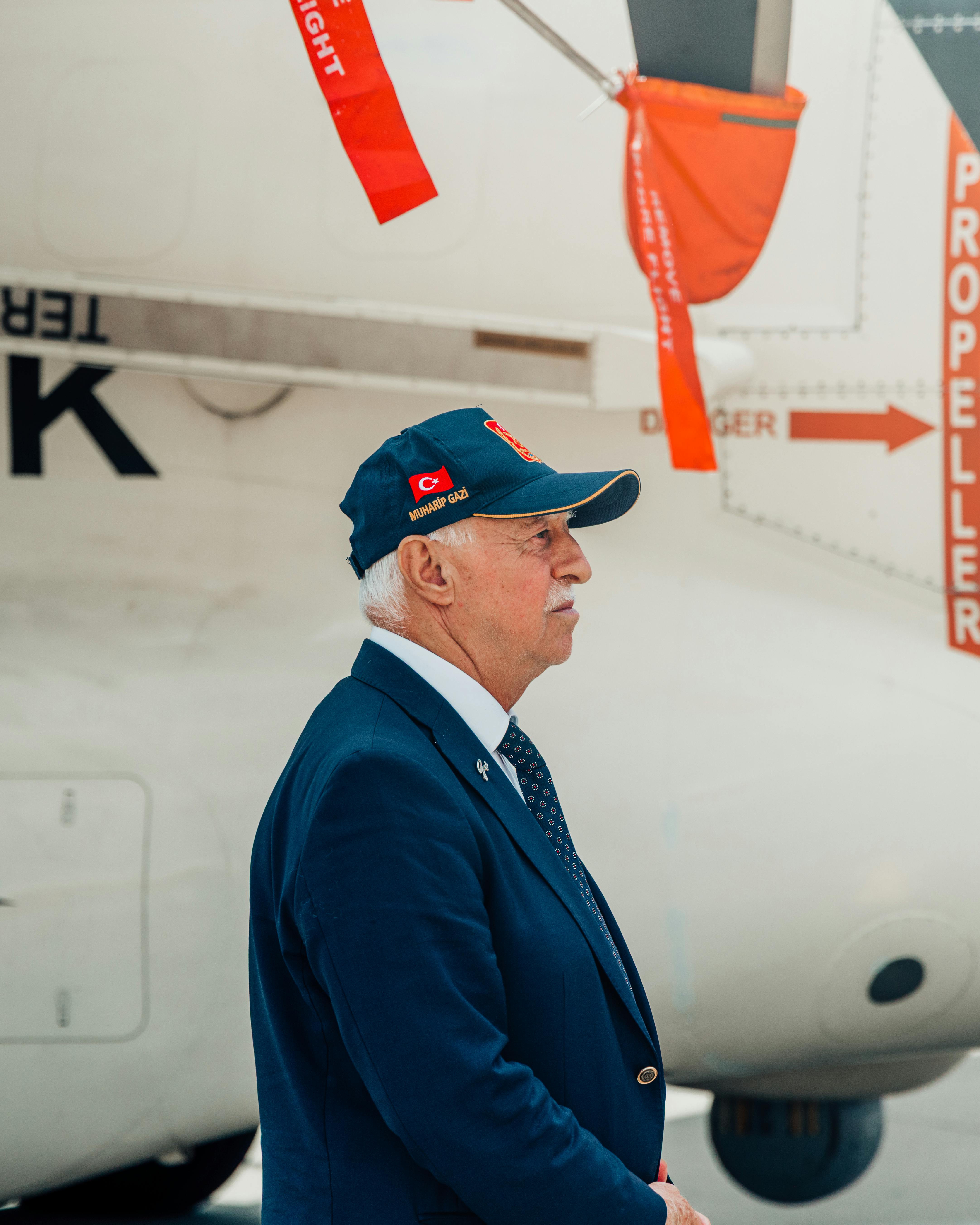 Elderly man wearing cap near airplane with visible propeller danger warning signs.