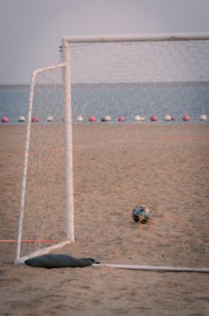 A soccer ball near a goal on a sandy beach by the sea at sunset, evoking a relaxed sporting atmosphere.