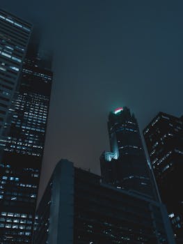 Dramatic nighttime view of towering skyscrapers in a foggy urban setting.