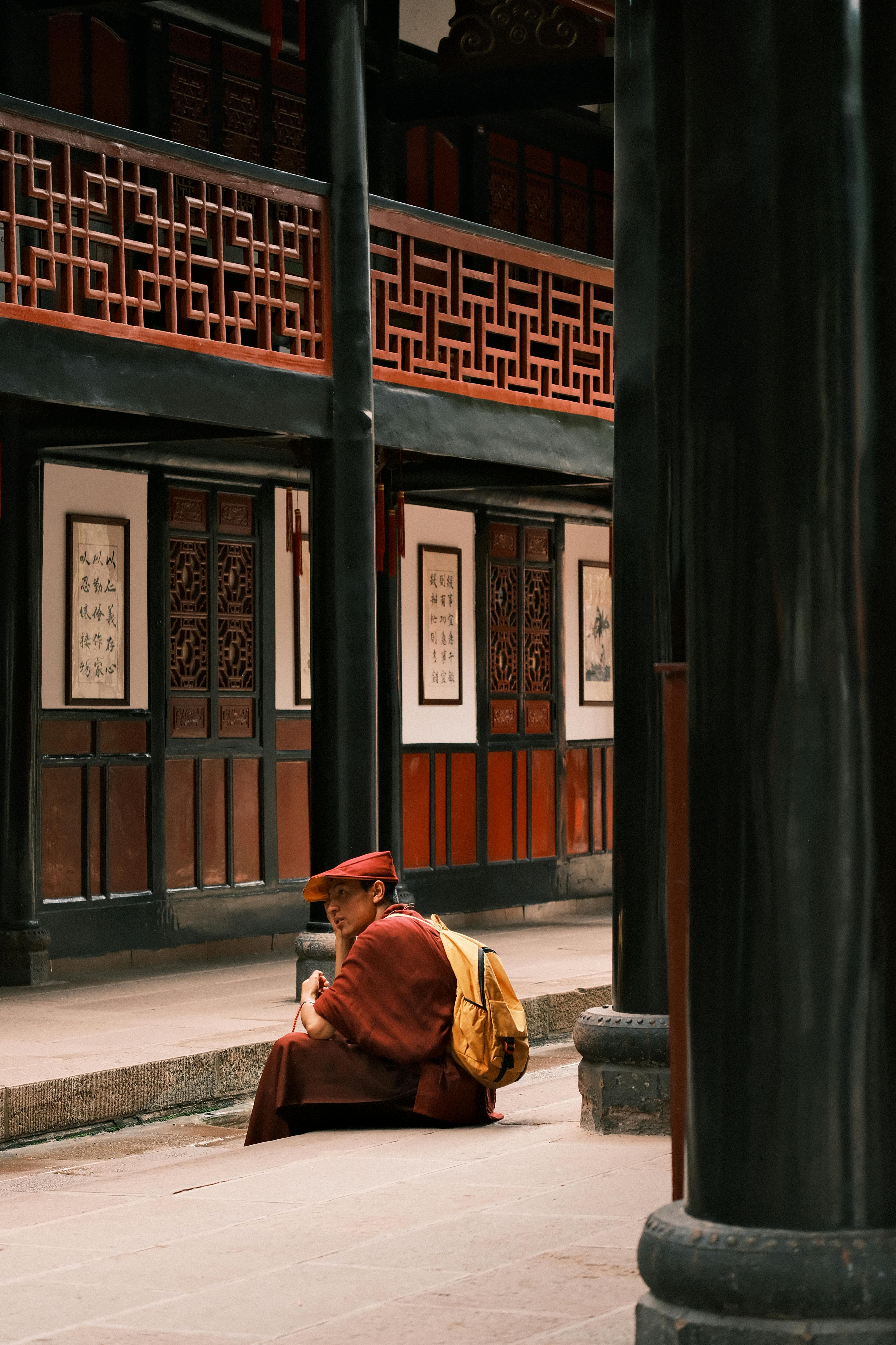 De franc Un monjo amb túnica vermella i motxilla groga seu tranquil·lament en un pati tradicional asiàtic, envoltat d'intricats dissenys de fusta. Foto d'estoc