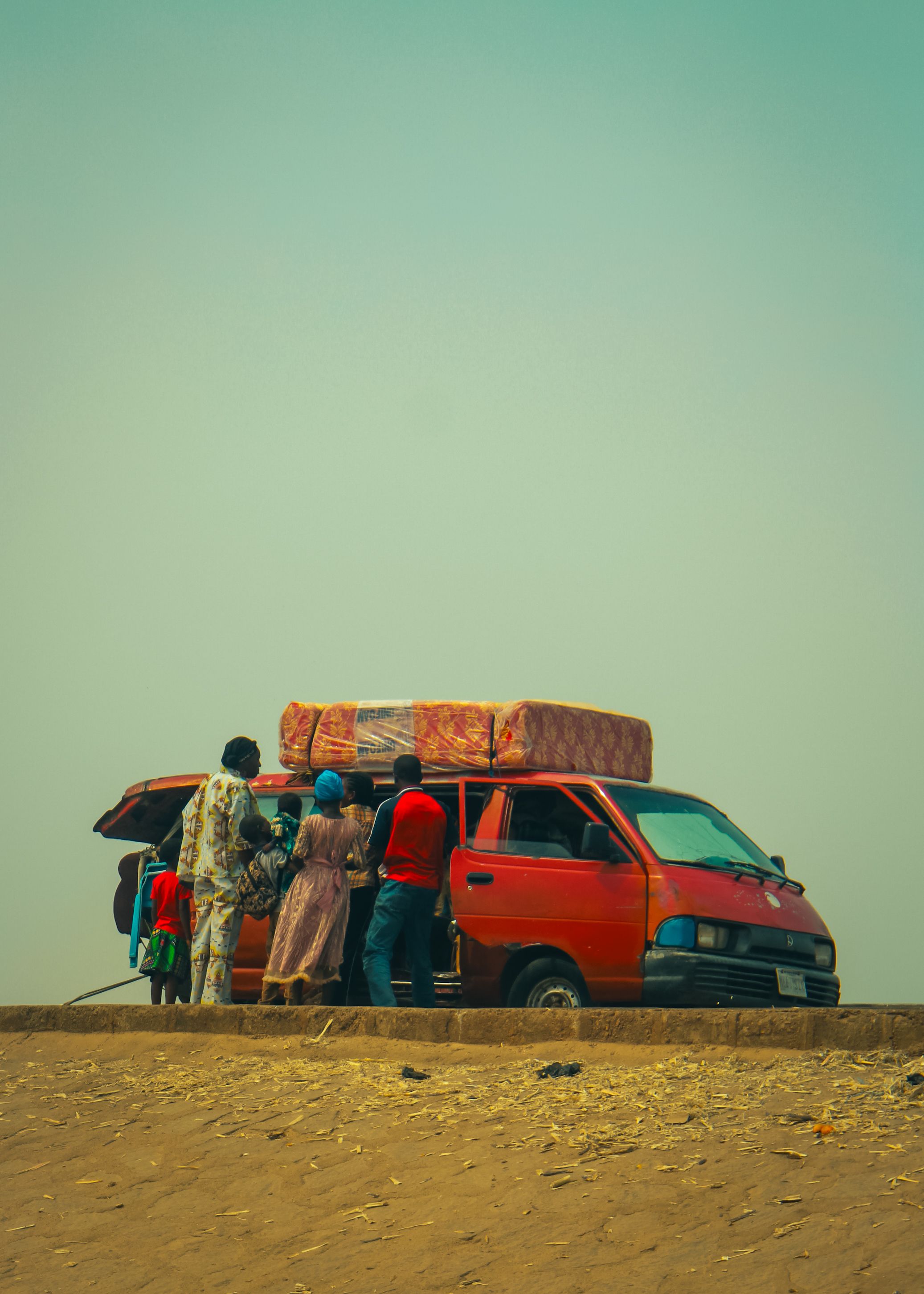A family gathers around a red van in a desert landscape, preparing for a journey.