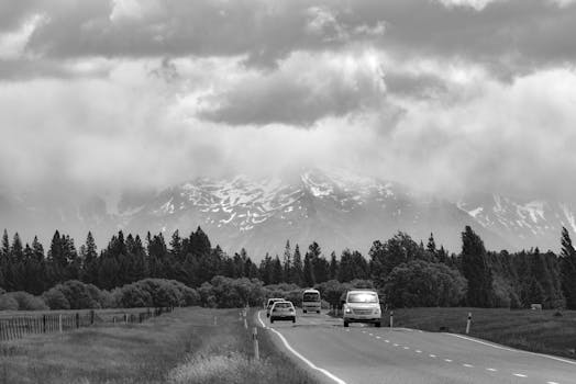 Scenic black and white view of cars traveling on a highway with dramatic mountains and clouds.
