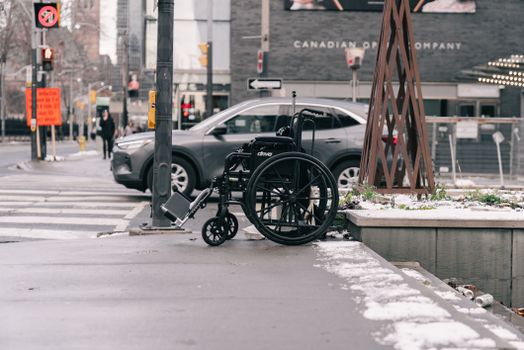 A wheelchair is parked on a snowy city sidewalk during winter with a car passing by.