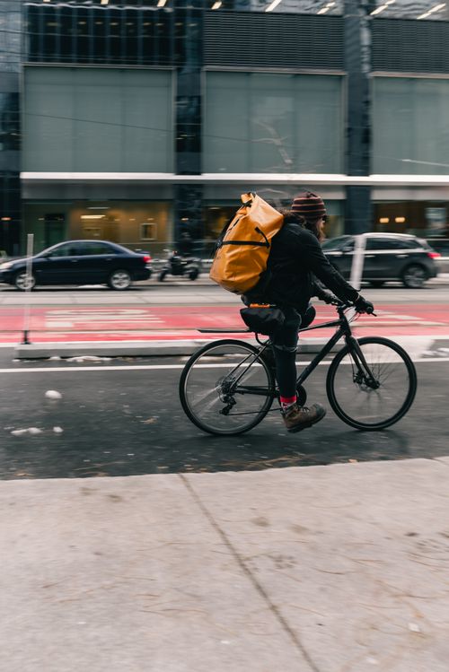 Free Cyclist with orange backpack riding in urban street, depicting modern city commuting lifestyle. Stock Photo
