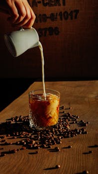 A rustic scene with milk poured into a glass of iced coffee, surrounded by coffee beans.