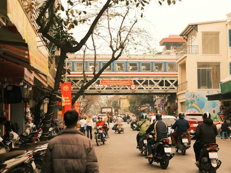 Vibrant street scene in Asia with people, motorbikes, and an overhead train.