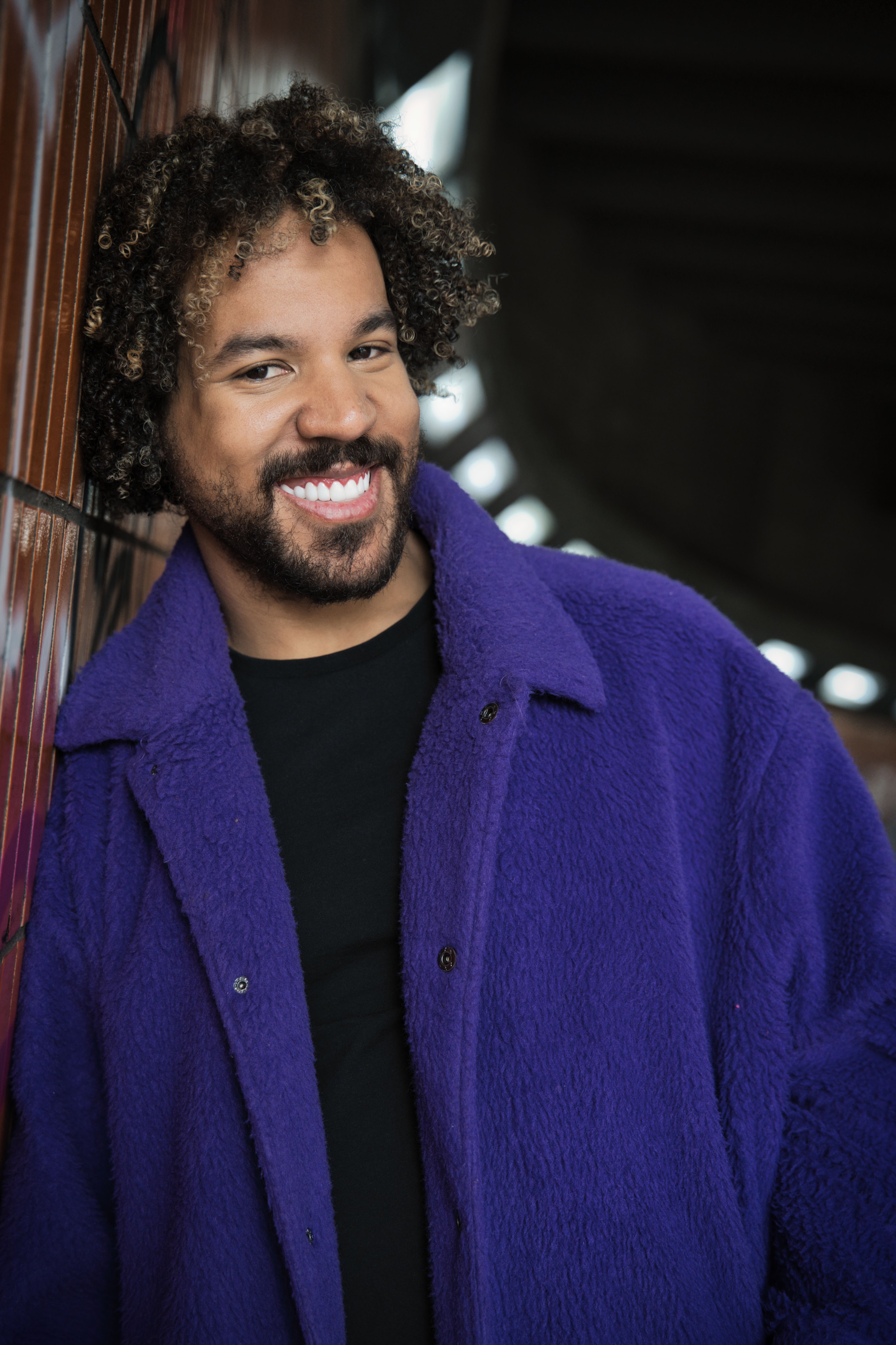 Cheerful adult man in a purple coat smiling against a graffiti wall in Berlin.