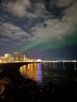 Stunning view of northern lights over Reykjavik skyscrapers and sea at night.