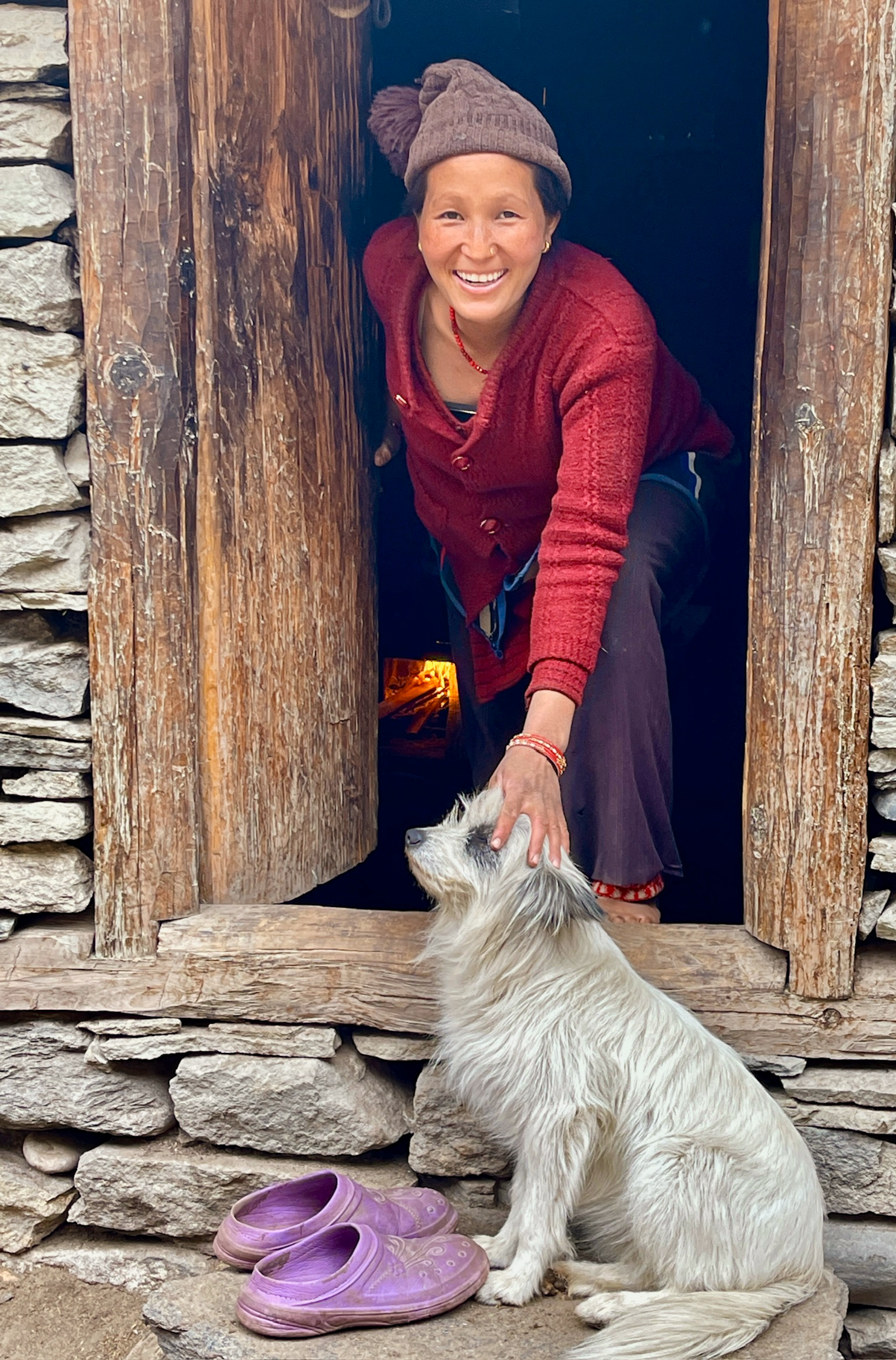 A cheerful Nepali woman pets a dog at a doorway in Karnali Province, Nepal.