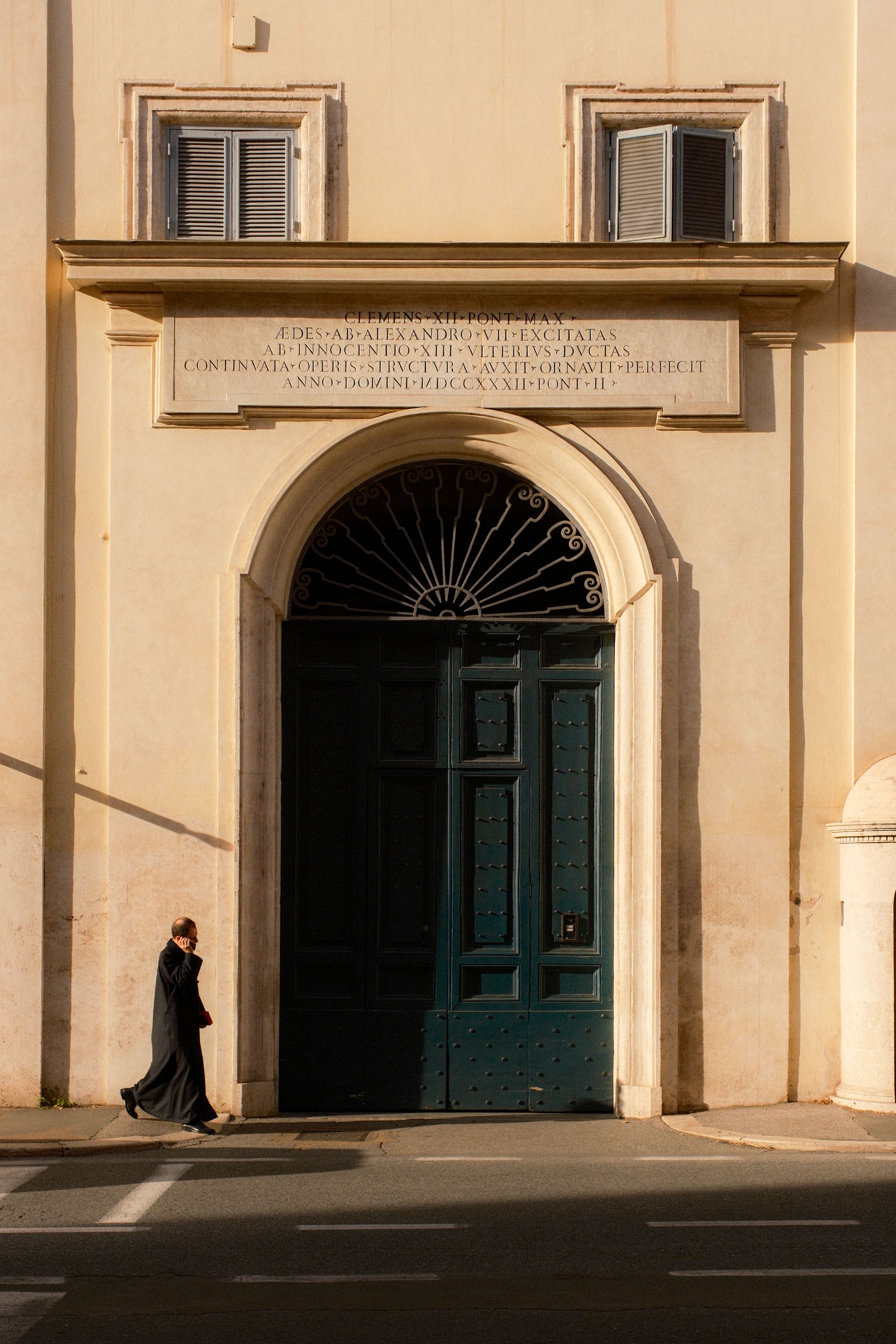 A lone person in black walks by a large ornate door on a sunny day.